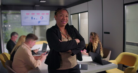 Smiling African American businesswoman standing with arms crossed in office, confident leadership presence as team collaborates in background with data screen
