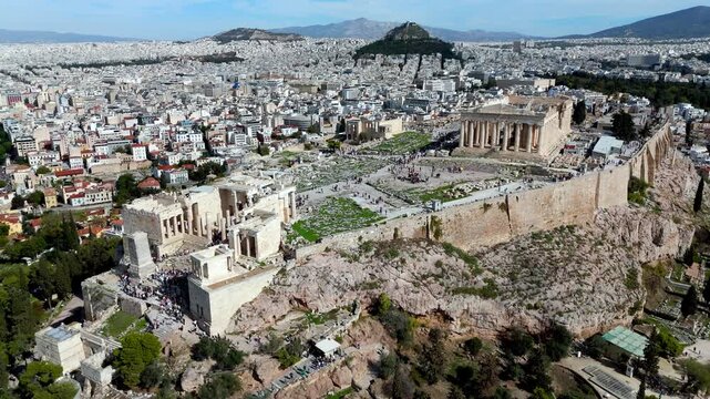 Flying high with zoom-out and lateral move over the Acropolis summit, revealing the Parthenon, Erechtheion, Athena Nike temple, tourists, and Lycabettus Hill.