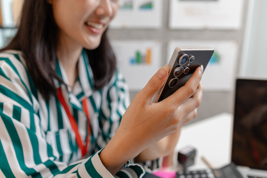 A young businesswoman is sitting in a modern office environment while holding her smartphone, symbolizing digital communication, business connection, and the spirit of entrepreneurship.