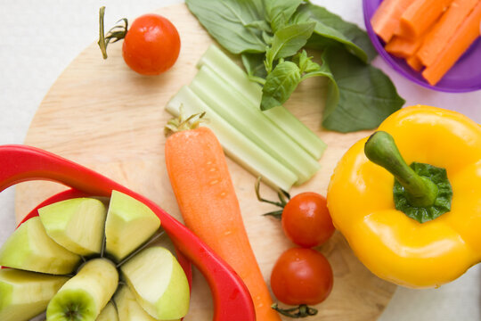 Fresh vegetables on a wooden cutting board: cherry tomatoes, celery sticks, carrot, yellow bell pepper, basil leaves, and sliced apple in a corer. Vibrant, healthy food preparation scene