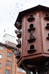 A wooden pigeon coop with several pigeons perched. A flock of birds takes flight in the background. Set against a brick building, this scene captures urban wildlife and avian life in harmony.