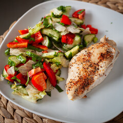 A healthy meal featuring a seasoned grilled chicken breast paired with a fresh vegetable salad. Crisp lettuce, juicy tomatoes, cucumber slices, and herbs create a vibrant, nutritious dish on a plate