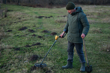 A man in a green jacket scans the ground with a metal detector while standing in a grassy field covered in small dirt mounds during early morning light