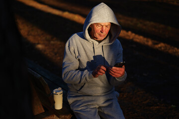 An elderly man sits on a bench in a park wearing a gray hoodie. He is focused on his phone as the warm sunlight casts long shadows around him