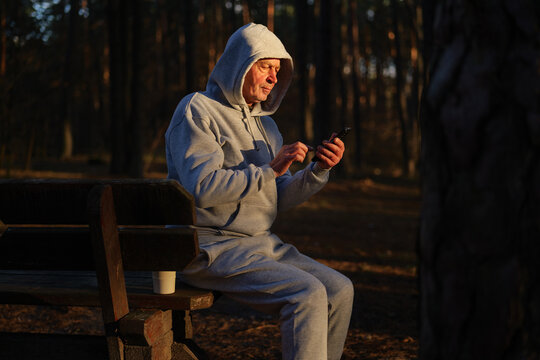 A senior man dressed in a gray sweatsuit interacts with his smartphone while seated on a bench in a quiet forest during the late afternoon light