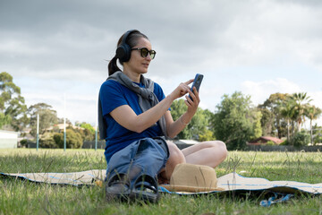 Woman relaxing in park wearing headphones and browsing on a smartphone.