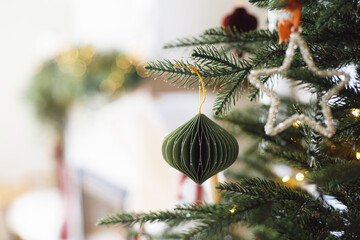 Stylish paper bauble on Christmas tree branches close up. Modern ornaments on decorated xmas tree with lights. Winter holiday banner, atmospheric christmas time. Merry Christmas!