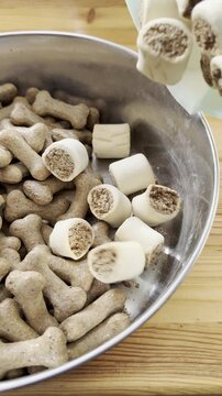 Vertical video social media - Closeup of dog biscuit cylinder shape rolls being poured into a large steel bowl, filling it up next to crunchy bone shaped biscuits.