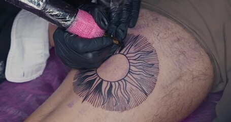Close-up of a tattoo machine needle injecting ink into the skin, outlining a sun design on a leg