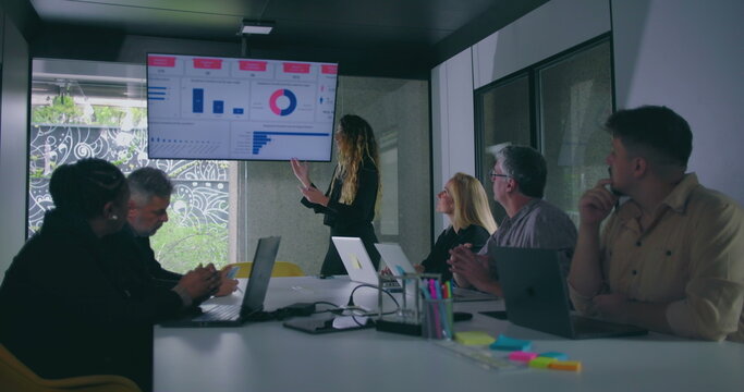 Team listens attentively as young woman presents data on wall screen during business meeting in modern office with diverse colleagues using laptops around table - Powered by Adobe