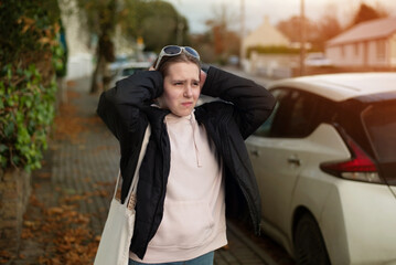 A young girl does not like the noise of big cities, she stands near road where there are many cars and covers her ears and looks anxiously ahead, her head starts to hurt