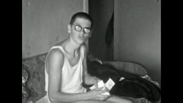 Young man with glasses sitting on sofa, looking through stack of old photographs, turning to look at camera with playful expression. Soviet house. Old film. Archival retro 1960s. Vintage archive.