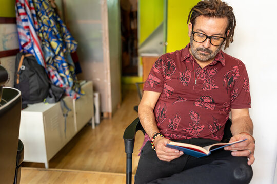 Mature Latino man with dreadlocks reading a book, waiting in a barber shop - Powered by Adobe