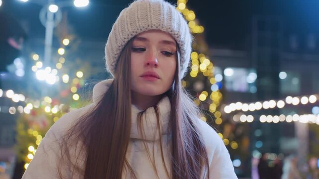 Young woman in warm winter clothing sits outdoors at night, appearing sad and contemplative, city lights glow around her as people skate, socialize, and celebrate