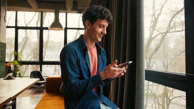 Smiling young man typing on smartphone using mobile app in modern apartment