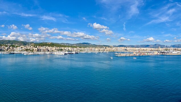 Scenic panoramic view of a coastal city with marina, yachts, and calm blue sea under a bright sky with scattered clouds, surrounded by hills and buildings