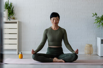 Woman practicing yoga meditation in lotus pose on mat at home, relaxing and focusing on mindfulness...