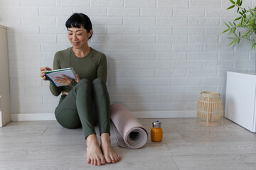 Woman in sportswear sitting on floor by yoga mat, writing in notebook and reflecting after home...