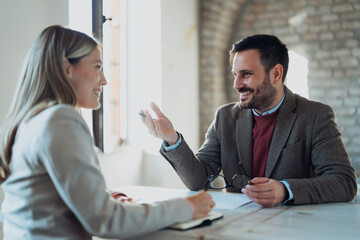 A professional man and woman sit across from each other at a table during a business meeting. The man, wearing glasses and a suit jacket, gestures with his hands while explaining a point.