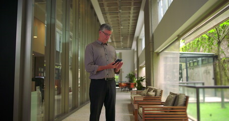 Wide shot of middle aged man using cellphone while standing in modern office hallway calm and focused posture interacting with mobile device in business setting