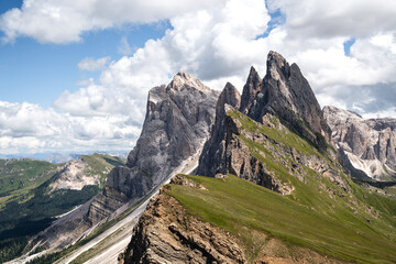 Dramatic view of the Odle peaks from Seceda in Val Gardena, Dolomites. Sharp limestone spires rise above green alpine ridges under bright blue sky with clouds an iconic landscape of the Italian Alps