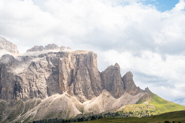 Dramatic view of the Torri di Sella peaks in Val Gardena, Dolomites. Sharp limestone spires rise above green alpine ridges under bright blue sky with clouds an iconic landscape of the Italian Alps