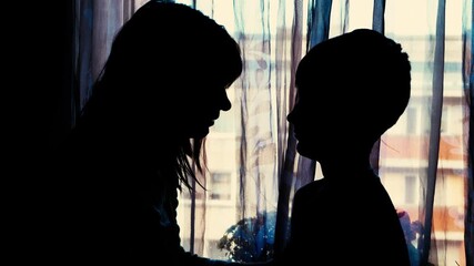 Two individuals are shown in silhouette near a window with sheer curtains; one appears to comfort the other in a moment of emotional support or tension.