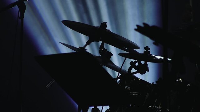 A drum kit is seen in dramatic silhouette with blue stage lights streaming behind it, creating a moody and atmospheric concert scene with copy space.