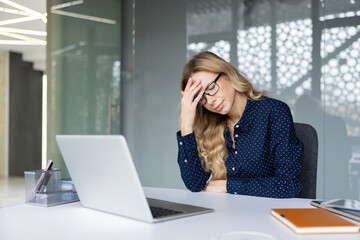 Businesswoman rubs her temples at a cluttered desk, exhausted and overwhelmed by deadlines and long hours, showing burnout, headache and workplace stress
