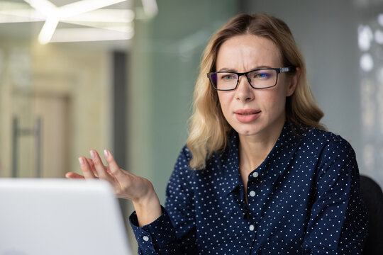 Young businesswoman with glasses experiencing confusion and frustration while having a video call on a laptop, gesturing during a remote meeting or online conversation in an office setting