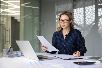 Businesswoman reviewing documents and data at her office desk, managing paperwork, and preparing financial reports with a laptop and calculator, representing professional efficiency and organization