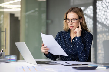 Fototapeta premium Young businesswoman wearing glasses focusing on financial documents while sitting at her desk with a laptop, clipboard, and pens in a contemporary office environment