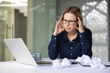 Businesswoman with glasses suffering from a headache and work related stress. Sitting at her desk with a laptop and crumpled papers. Feeling overwhelmed and exhausted in her modern office environment