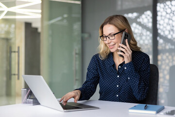 Blonde businesswoman talking on a mobile phone while typing on a laptop, effectively managing multiple tasks and communicating professionally in a bright modern office workspace
