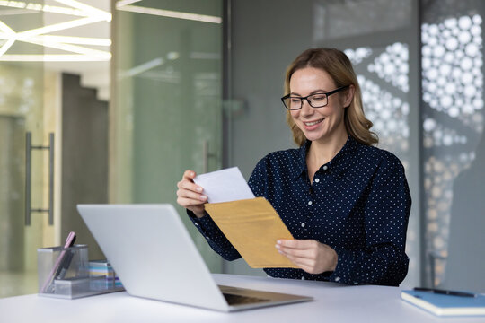 Happy businesswoman in glasses and polka dot shirt pulls a letter from a brown envelope at her office desk, smiling at a job offer or positive work-related news while laptop rests nearby