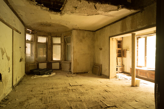 Sunlit abandoned interior with peeling plaster, broken ceiling and damaged wooden floor - dilapidated bay-window room for renovation or atmospheric background
