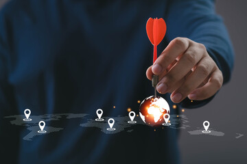 A hand holding a red dart targeting a glowing world map with location markers, symbolizing global business strategy, market targeting, planning, direction, and international growth opportunities.