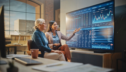 Financial Analysis Meeting Two Businesswomen Reviewing Stock Market Charts on a Large Screen Display in Modern Office Setting.