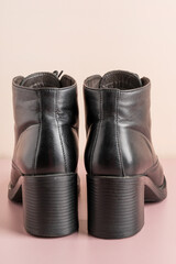 Close up rear view of pair of female ankle boots hand-sewn of shiny natural black leather with block heels and sole on pastel beige background used as warm casual comfortable season classic footwear