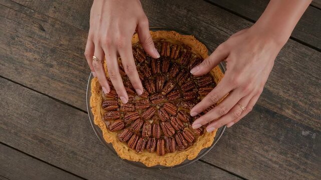 Entering hands adjusting pecan pie in glass dish on rustic tabletop before serving, wearing rings