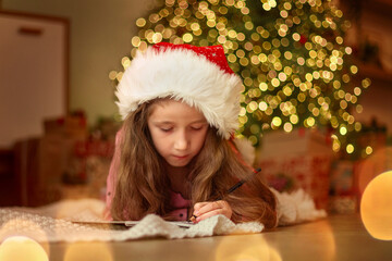 Little girl in red hat  writes a letter to Santa Claus near the Christmas tree