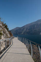 Obraz premium Panoramic view of the lakeside walkway in Limone sul Garda, curving along the cliffs above the water and offering a scenic route with bright reflections and dramatic mountain backdrops.