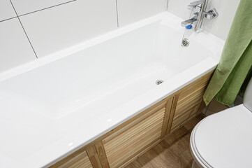 A clean, white bathtub with wooden paneling is seen next to a toilet. A green towel hangs nearby, set against white tiled walls and a wooden floor
