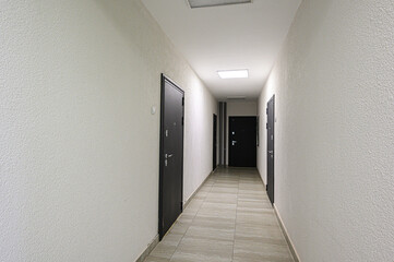 A well-lit corridor features textured white walls and dark doors, with tiled flooring leading deeper into the space under evenly spaced ceiling lights