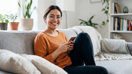 Young Indian woman sitting comfortably on couch browsing phone indoors