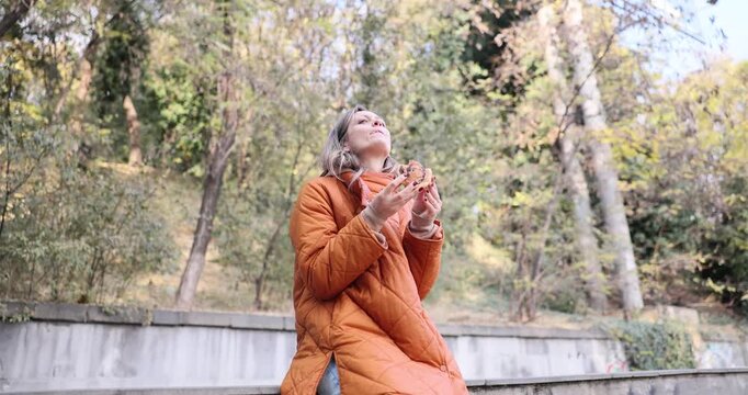 Woman sits on parapet outdoors in park area holding warm pastry. Female bites fresh bun enjoying quick snack during calm city walk slow motion