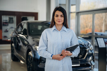 Notepad is in hands. Woman posing with a car in an auto showroom, modern dealership environment