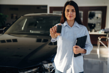 Daytime, natural lighting, holding keys. Woman posing with a car in an auto showroom, modern...