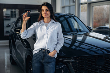 Daytime, natural lighting, holding keys. Woman posing with a car in an auto showroom, modern dealership environment