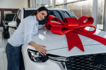 Red gift ribbon on a car. Woman posing with a car in an auto showroom, modern dealership environment